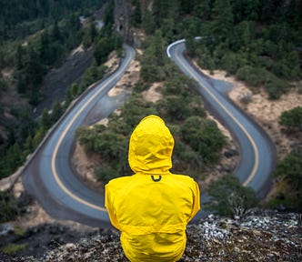 Woman in a yellow rain slicker staring at a wet black crossroads, a fork in the road. She has a choice to make.