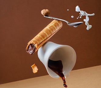 A photograph freezes a cup of coffee spilling forward, with a cinnamon stick just above it, a spoon above the cinnamon stick, and a splash of milk above the spoon. Sugar and fat intake affects the gut microbiome.