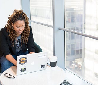 A Black woman sits next to a big window and types on her computer. Her glasses and coffee are sitting nearby.
