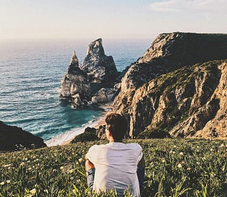 A man sits on a grassy hillside, gazing at a stunning coastal view with rocky cliffs and the ocean in the background.