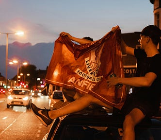 Two Liverpool Football Club fans sit on the roof of a car at the side of a city street, holding a club flag