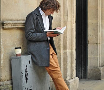 A man is leaning against a wall, reading a book.