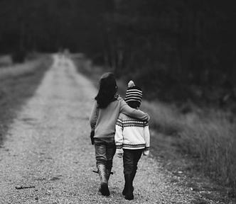 Two young children, one draping an arm over the other, both facing away from the camera, walking down a dirt/gravel road to a distance not established. Upon the sides of the road are brush that are bristlely, short, and dry grass.