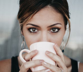 A Brunette with big brown eyes looking straight into the camera sipping coffee from a pink mug