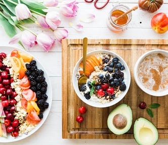 a photo of a platter or colorful fruits, a bowls of oatmeal with fruit, a sliced avocado, and a cup of coffee