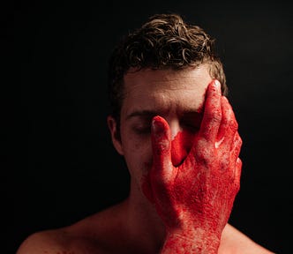 A middle-aged man’s face is almost covered by a blood-stained overly large hand not his own.