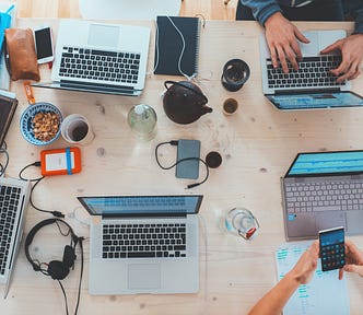 There are four laptops on a table, it’s an overhead shot. The group of people in the meeting might be engaging in a meeting about digital marketing. One person is holding a smartphone with some papers with green handwriting on the paper. There are one pair of black headphones, a glass of water, a bottle of water, a teapot, a cup of coffee, a bowl of cereal in a white and blue bowl, some pens, an orange hard drive, some notebooks, smartphones and another grey hard drive on a pale colored desk.