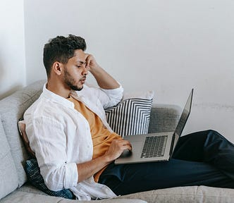 Man in a white shirt sits on a couch looking at his laptop.