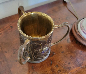An old looking pewter cup standing on a wood-grain table. The cup has three handles sticking out from it.