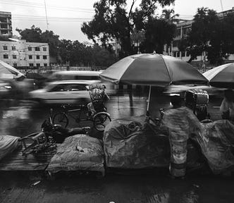 A bustling street comes to life in the rain — vehicles speeding, rickshaws weaving through traffic, and vendors preparing to sell fresh produce. The wet roads reflect the hurried pace of the city, mirroring the endless chase for more. Yet, in the rush to move forward, are we missing the beauty of the present? — alt text to boost Medium stories.