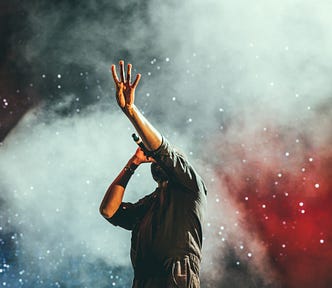 A music artist on stage reaching up with one hand while they sing. The crowd is shining lights in the background, and a white smoke envelopes the scene.