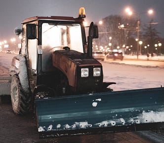 Small snow plow at night. City lights in the background.