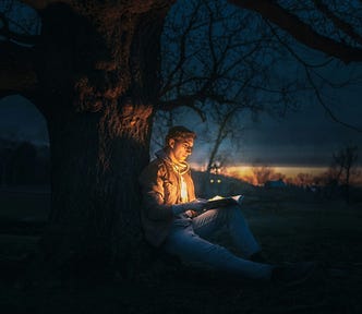 A man sitting beneath a tree at night, reading a book. The book shines a golden light.