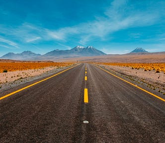 Two-lane paved road heading toward mountain horizon with blue sky.