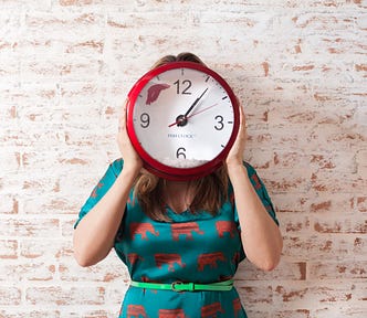 A woman faces us, holding her arms up to hold a face-concealing clock. The time device has a red rim, white face, and black hands. The time is 1:06. White brick wall in background. The woman dons a blue dress with repeated red elephants across it.