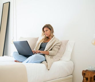 A blonde woman is sitting in bed, typing on her laptop.