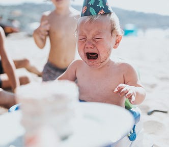 A toddler on the beach sitting in a high chair is crying and wearing a party hat. An older boy looks on.