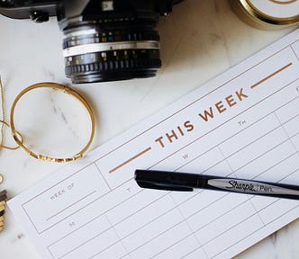 Weekly planner on busy desk with a pen, a necklace, a camera and tape.