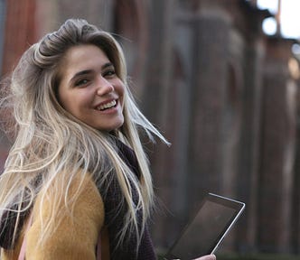 Confident blond, smiling woman looking back at us over her shoulder while holding an i-Pad. She’s outside and wearing a thick wooly jumper