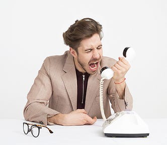 Young man in beige blazer sitting at a desk screaming into an old fashion white phone