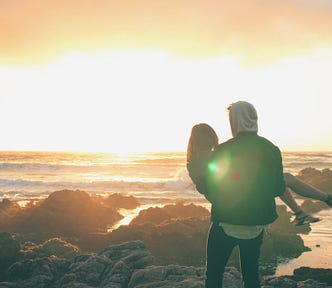 A romantic photo of a man carrying a woman. They’re both facing away from the camera and towards a yellow and orange sunset over a rocky beach. The light of the sun overexposes the photo, a light burst shining in one spot near his shoulder and hers. He supports her in his arms, in front of him. Her head is visible behind his left side, and her legs on his right. She’s wearing heels, which are not ideal for the rocky beach conditions.  They look out towards the sunset.