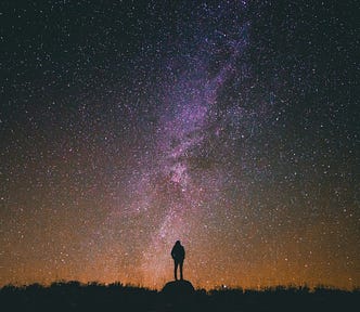 A lone figure stands under a vast, starry sky with the Milky Way stretching above, symbolizing the profound connection between humanity and the universe.