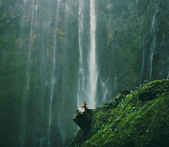 A huge waterfall behind the person who sits on a rock.
