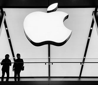 A large, illuminated Apple logo prominently displayed in a store. Below the logo, two individuals are standing at a balcony, likely within the Apple Store. The black and white tones enhance the modern and sleek aesthetic typical of Apple’s store design. This setting, with its sharp contrasts and iconic logo, effectively highlights the branding and architectural elements that Apple is known for.