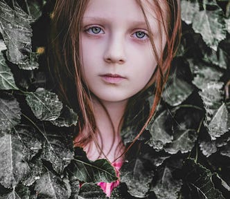 A little girl’s face emerges, while her body is partially hidden beneath a layer of leaves
