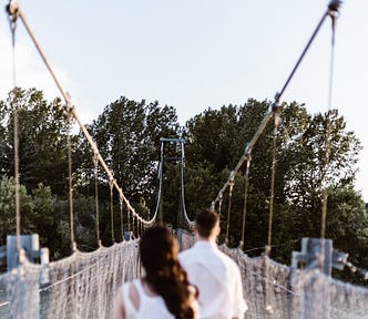 A couple walk away from the camera, beginning a journey across a bridge to a destination hidden by trees