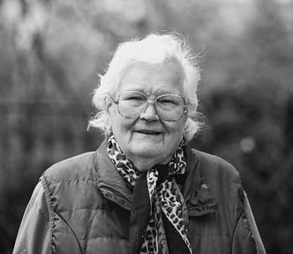 A black and white photo of an old lady with gray hair, glasses and a sly grin