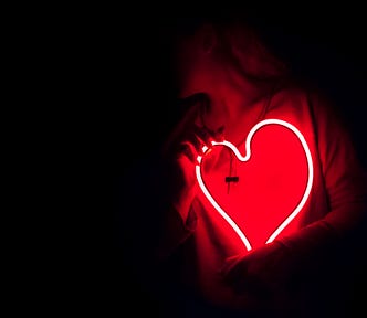 Black background with a woman’s silhouette holding a white neon heart in front of her red t-shirt