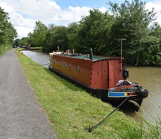A moored narrowboat on a canal with a broad towpath.