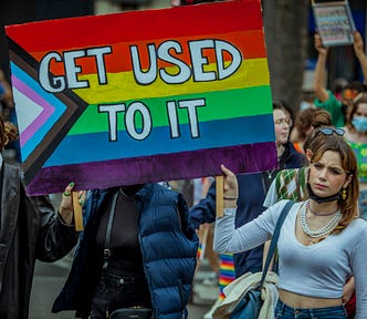 A Pride parade with several people marching. The two people in the foreground are holding a large Progress Pride flag on which is written in block letters, “GET USED TO IT.”