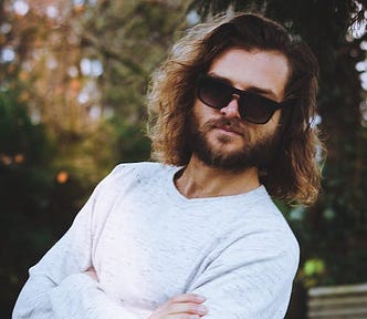 Bearded man wearing sunglasses with woods behind him