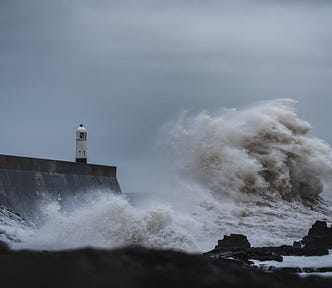 A huge wave crashes against a harbour wall with a small lighthouse