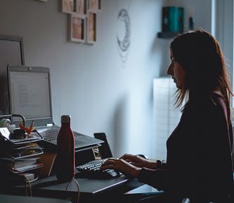 Woman at her desk, writing on laptop. With waterbottle, stationary, pens, paper etc