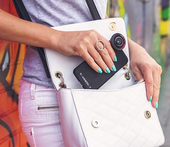 A white handbag is slung across a woman’s body. It is open and the woman is slipping her phone into it. She has turquoise nail polish on and behind her is a wall with red and green graffiti.
