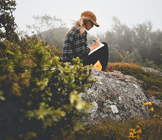 A woman with causal clothes and a cap, in the outdoors, sitting near a rock with small yellow flowers, writing in a notebook. It’s a cloudy day.