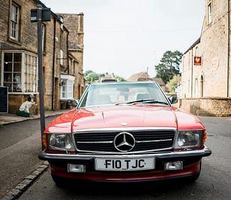 A red vintage Mercedes-Benz car is parked on a quaint street lined with stone buildings. The license plate reads “F10 TJC.” The background features a mix of charming shops and trees under a cloudy sky, adding to the picturesque, classic feel.