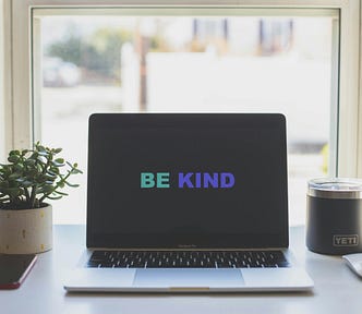 Laptop next to a window, plant, and mug. The computer screen says “be kind.”