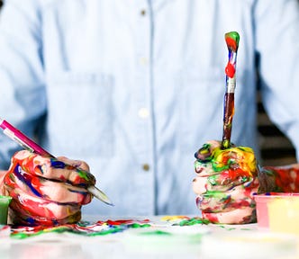 A picture of a person sitting at a table with their hands covered in paint. In one hand, they are holding a pencil. In the other hand, they are holding a paintbrush.