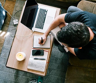 Man sitting on sofa with laptop on coffee table, notebooks, a phone and a candle, writing by hand.