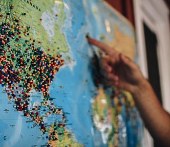 World map on cork board. USA and Central America covered in map tacks. Man’s finger pointing to one tack on Iceland.