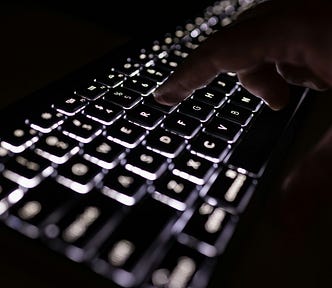 A backlit black computer keyboard in the dark, with the silhouette of someone pressing a key.
