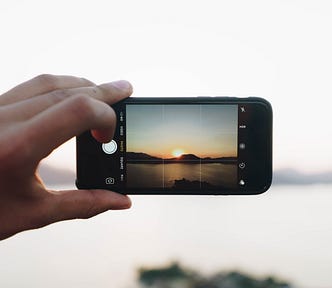 The cell phone camera is activated and a scene of a lake is on the screen while a hand is about to click the round button that takes the photo