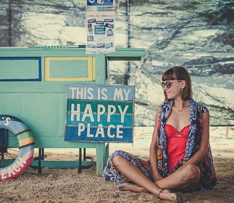 Women sitting on a beach, next to her is a wooden sign that reads “This is my happy place”