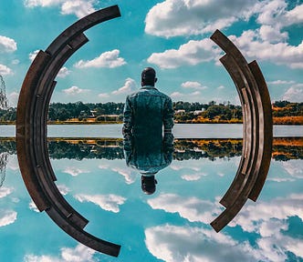 Man standing near a pond, facing away, the sky and his back reflected in the water