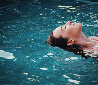 This image shows a person floating on their back in clear, blue water with their face pointing up to the sky. They have a look of contentment on their face. Only the person’s shoulders, head, and face are visible from the right side of the frame.