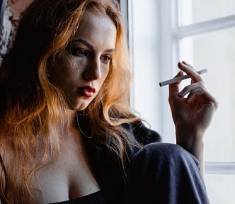 Photo of a light-skinned, copper-haired, distressed young woman wearing a black tank top, smoking a cigarette, in low light by a window.